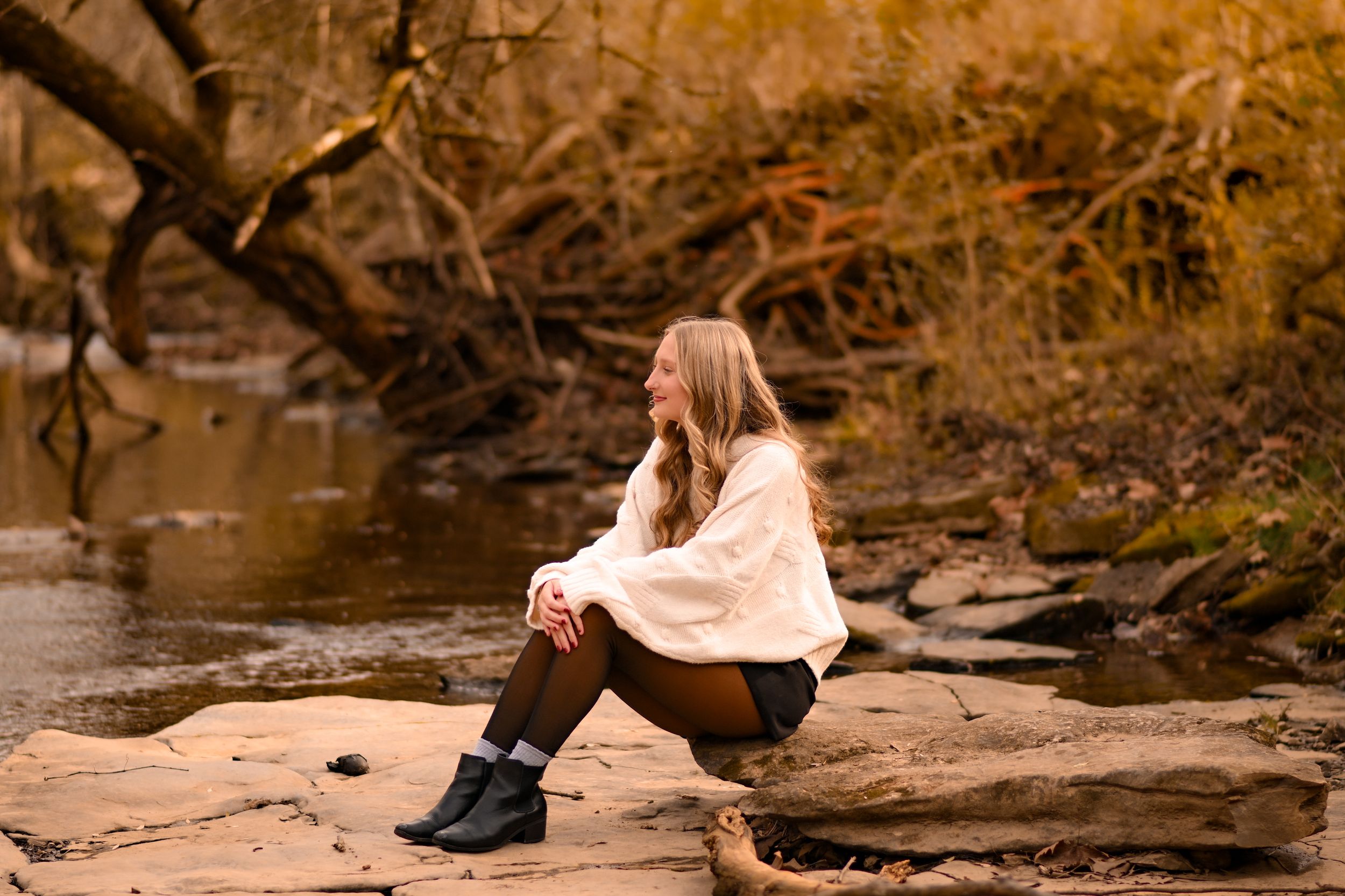 Woman in cream sweater sitting on rocks by stream during autumn.