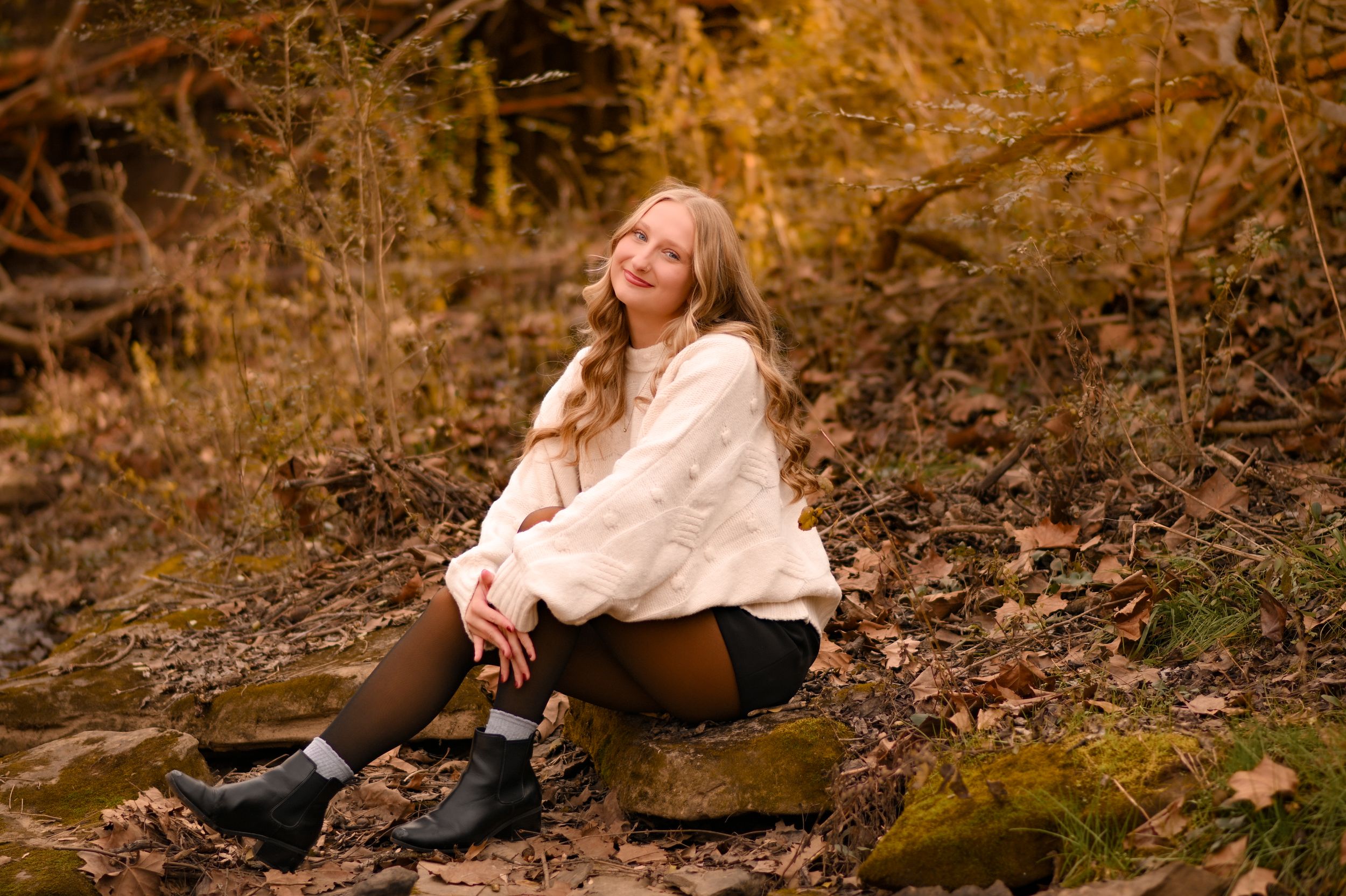 Woman in cozy sweater sitting on rocks surrounded by fall foliage.