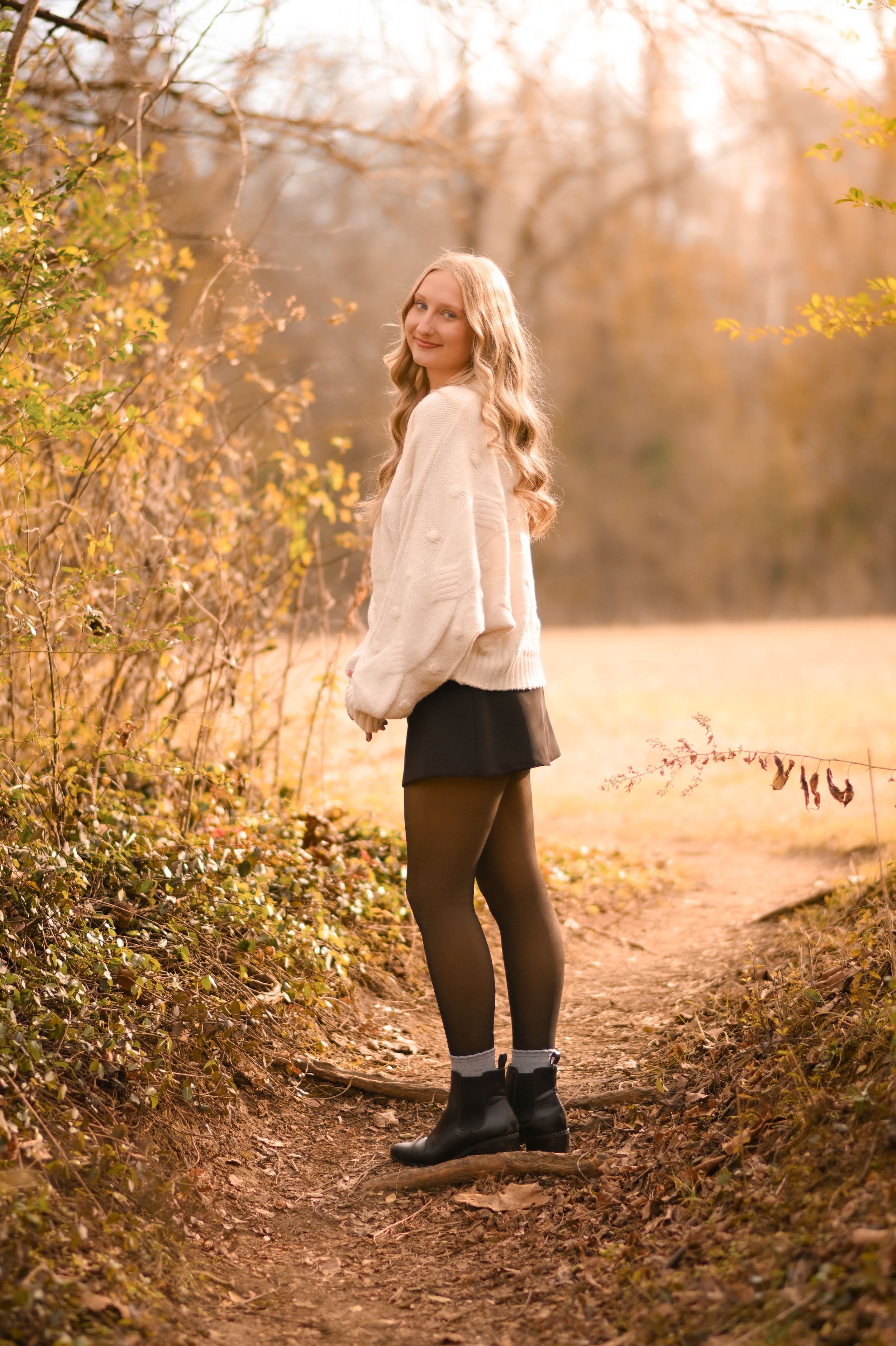 Woman in white sweater poses on autumn trail with golden sunlight filtering through trees.
