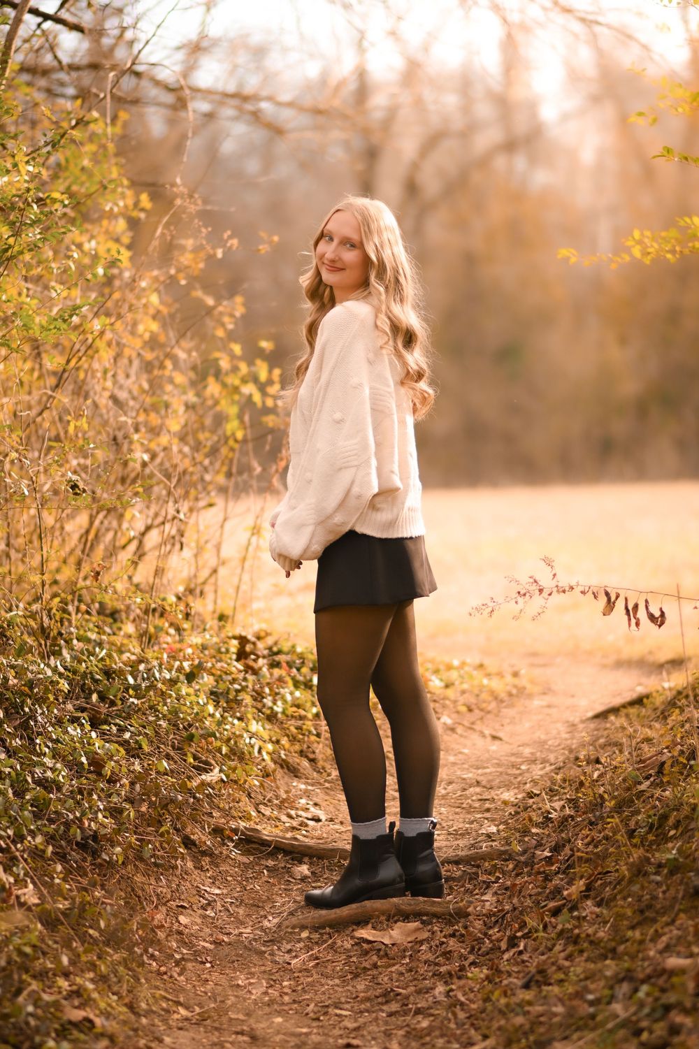 Woman in white sweater poses on autumn trail with golden sunlight filtering through trees.