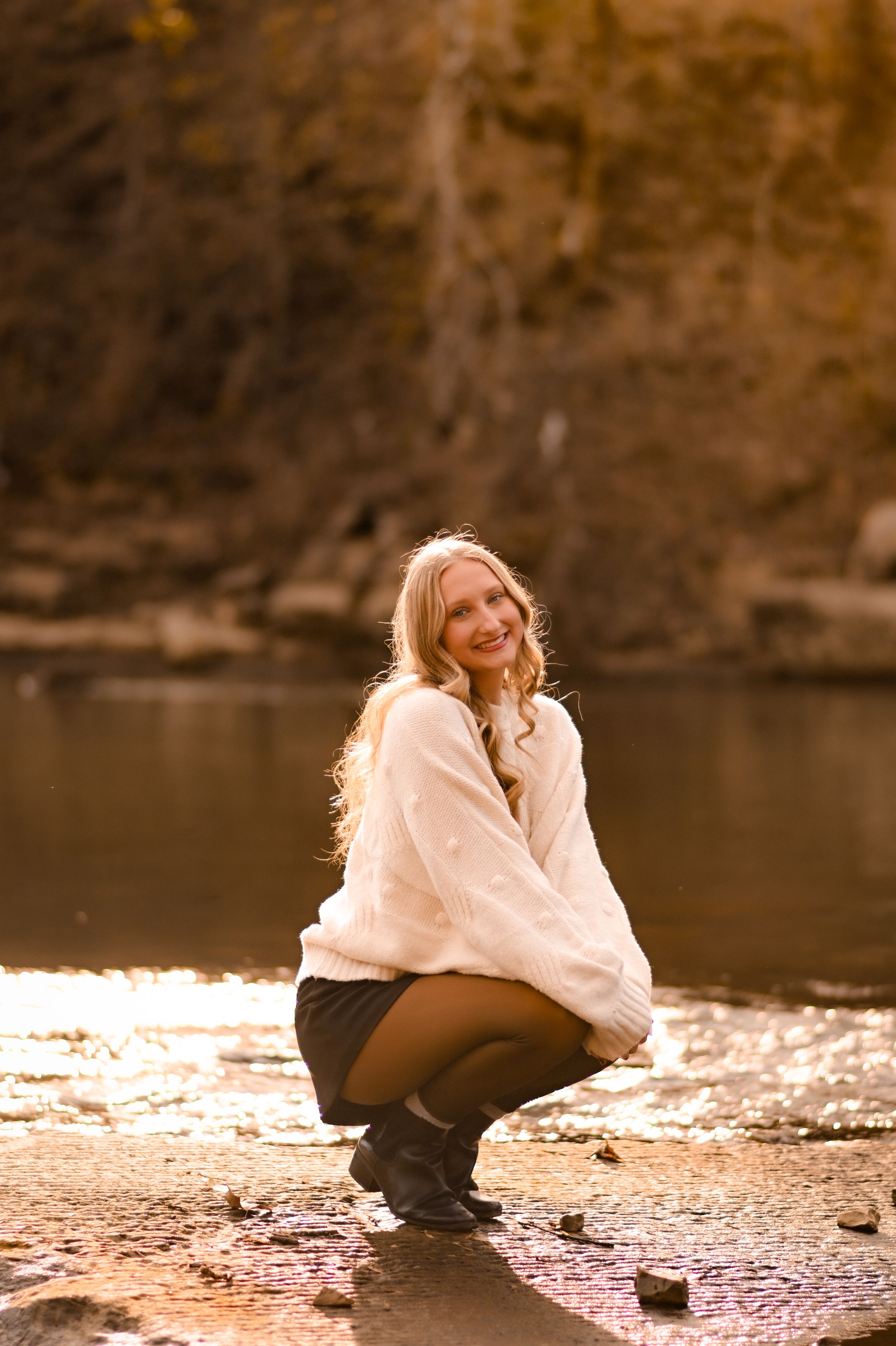 Woman in cream sweater crouching by water's edge during golden hour.