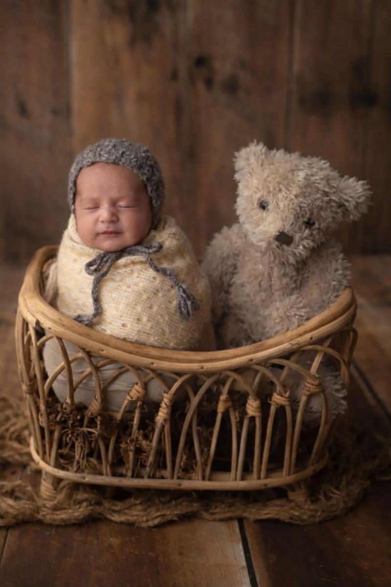 Sleeping newborn wrapped in burlap wearing gray knit bonnet sitting in wicker basket beside a teddy bear.