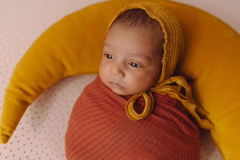 Newborn baby wrapped in rust swaddle wearing yellow knit bonnet, resting on crescent moon pillow.