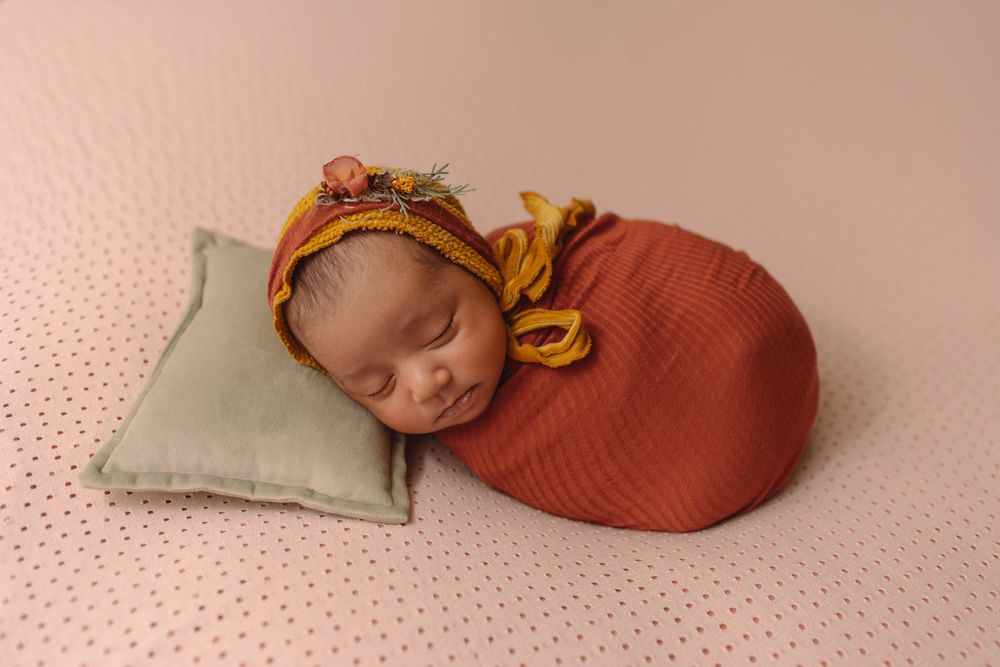 Newborn baby swaddled in rust wrap with floral bonnet, sleeping on mint pillow against pink backdrop.