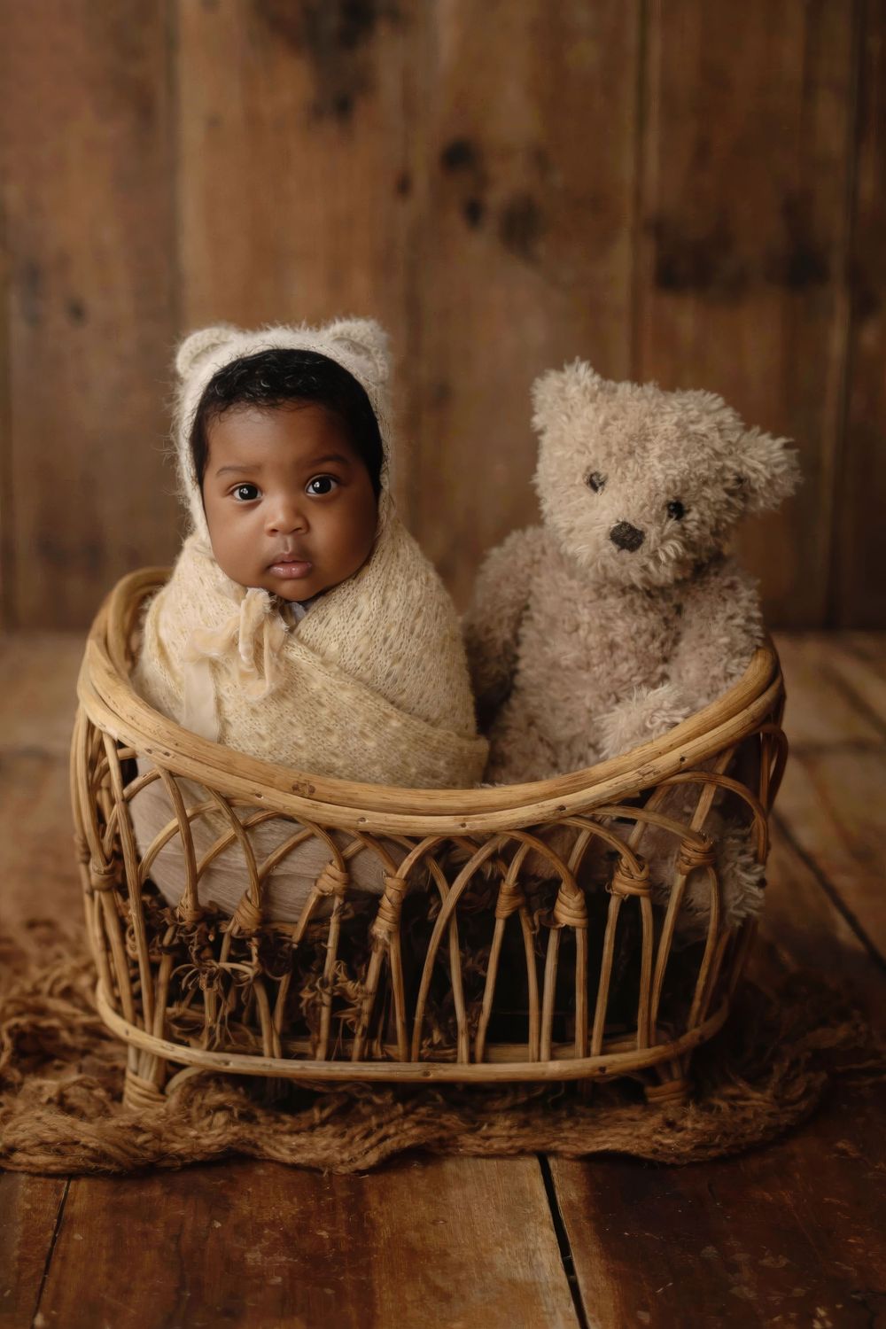 Newborn baby wrapped in cream knit swaddle wearing bear bonnet sits in wicker basket with teddy bear on wood floor.