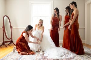 Bride in white gown with bridesmaids in rust velvet dresses helping with shoes in bright room with mirror.