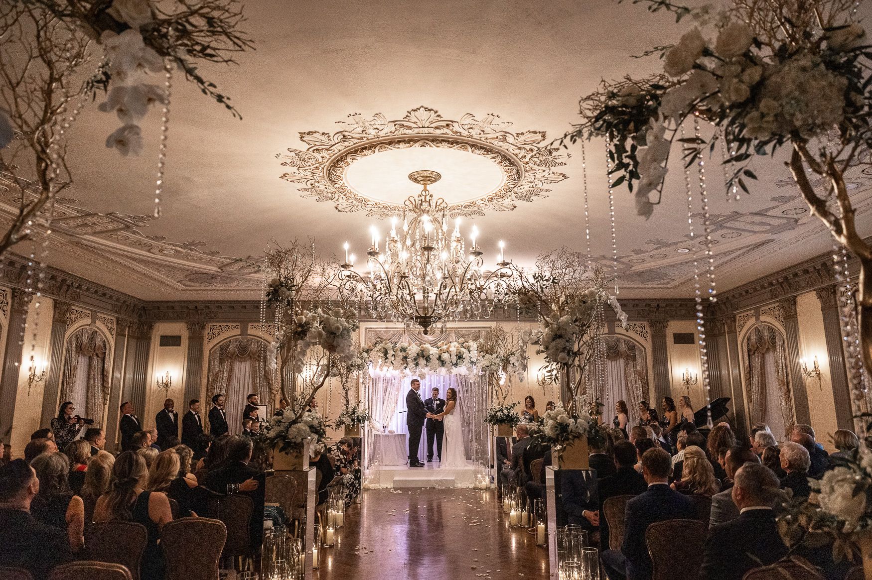 Elegant wedding ceremony in ornate ballroom with crystal chandelier, floral arrangements, and guests seated in candlelit venue.