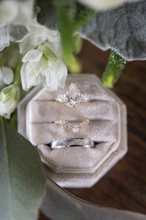 Three wedding rings displayed in a velvet octagonal ring box surrounded by white flowers and greenery.