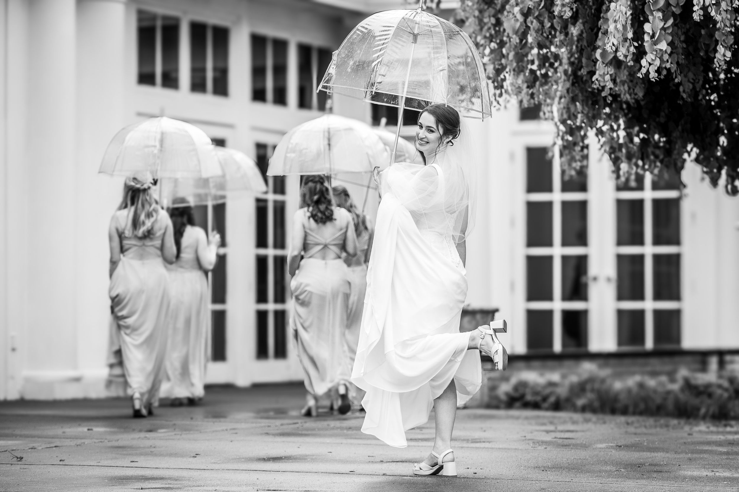 Bride and bridesmaids walking in rain holding clear bubble umbrellas in elegant white gowns, black and white wedding photo.