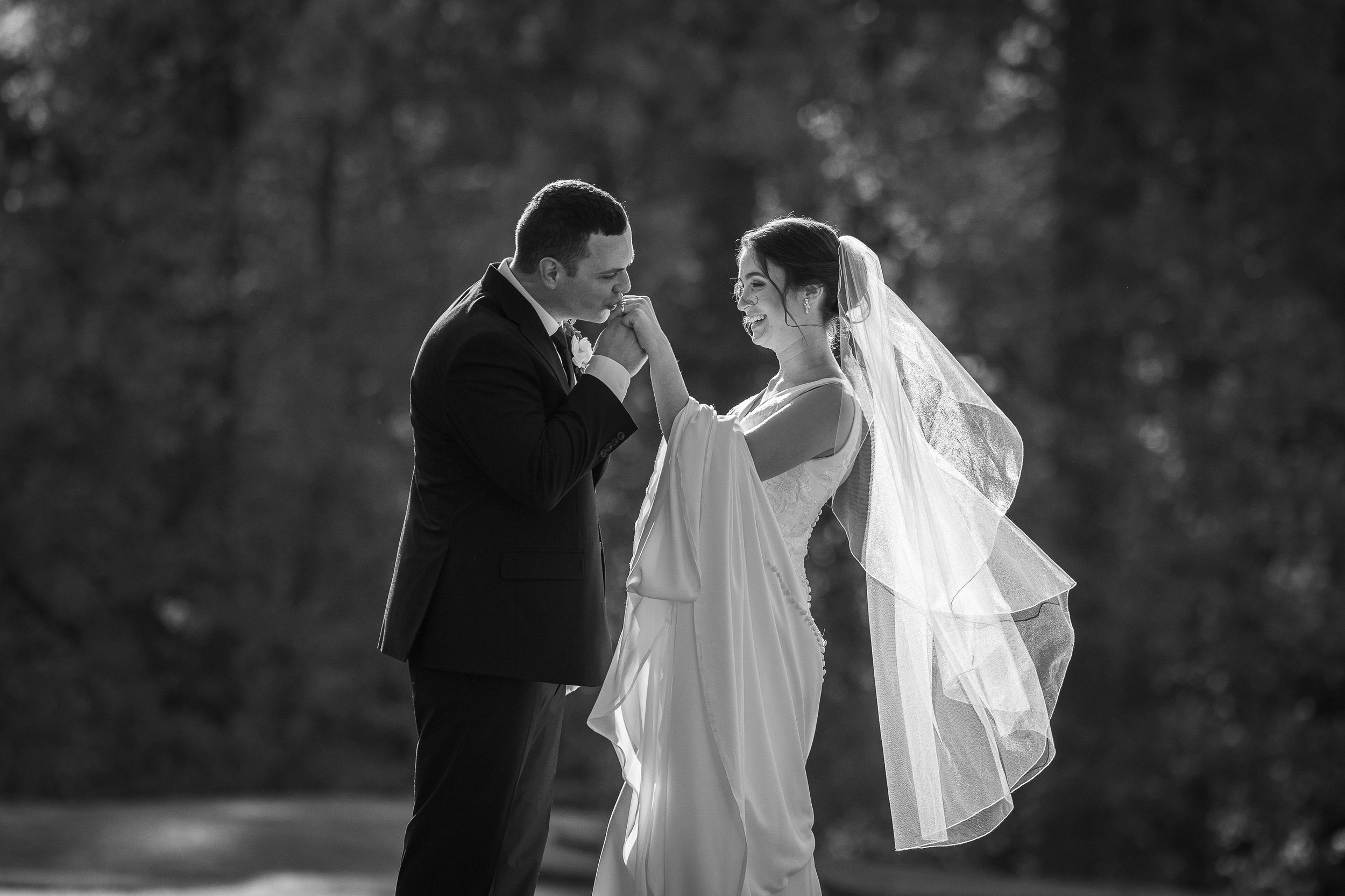 Black and white wedding photo of groom kissing bride's hand while she smiles, veil flowing, outdoors.