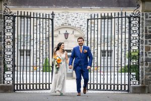 Bride and groom walking through ornate iron gates, groom in blue suit, bride holding colorful bouquet.