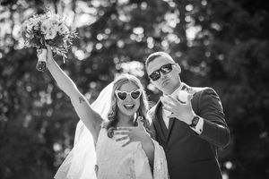 Black and white photo of a joyful bride and groom wearing heart sunglasses, celebrating their wedding day outdoors.