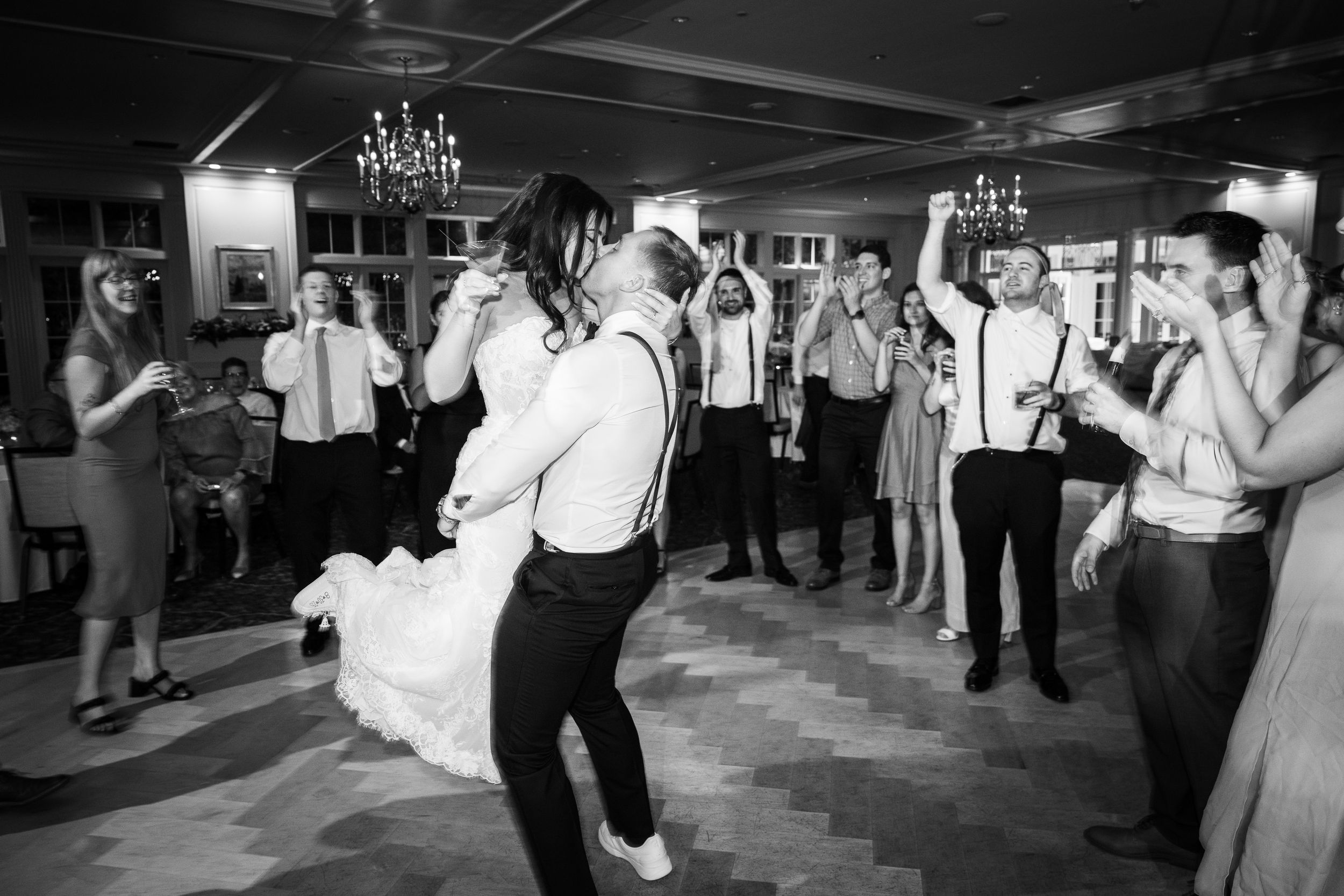 Black and white photo of bride and groom dancing joyfully at wedding reception surrounded by cheering guests.