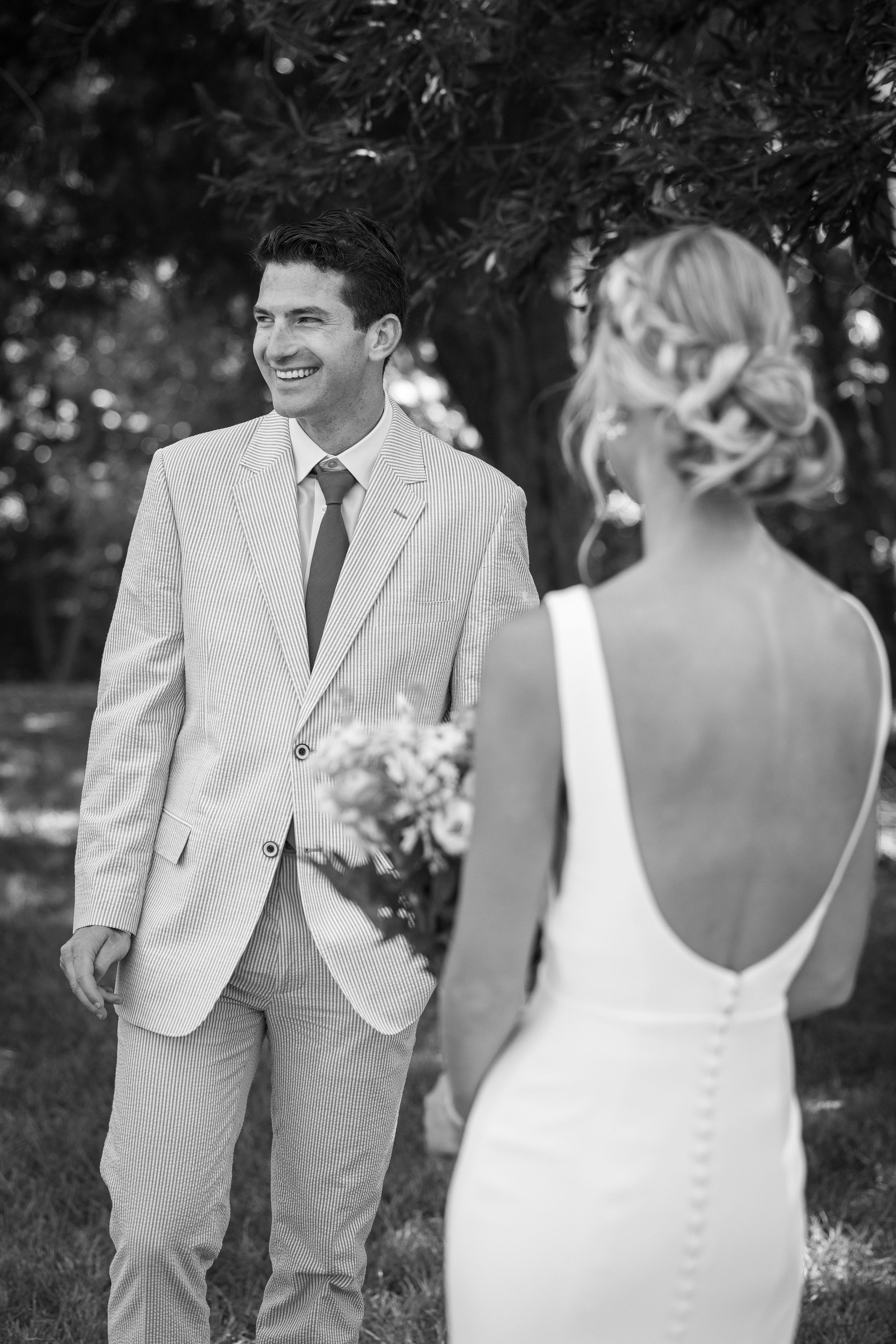 Black and white photo of smiling groom in light suit seeing bride in backless wedding dress for the first time outdoors.