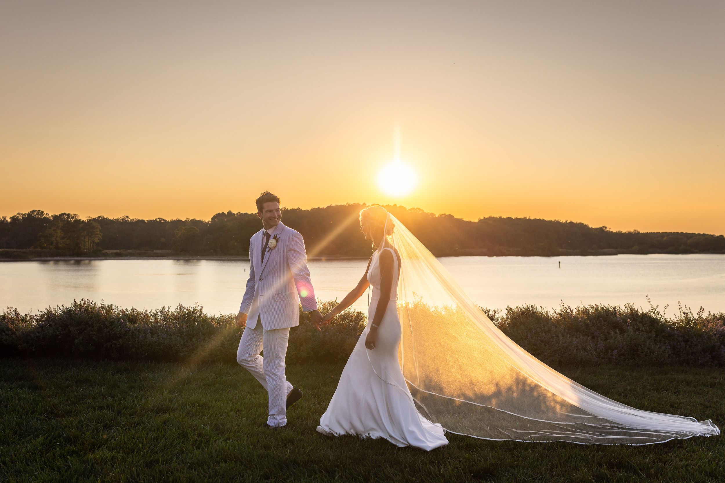 Bride and groom hold hands at sunset by a lake, her long veil flowing, in a romantic golden hour wedding photo.