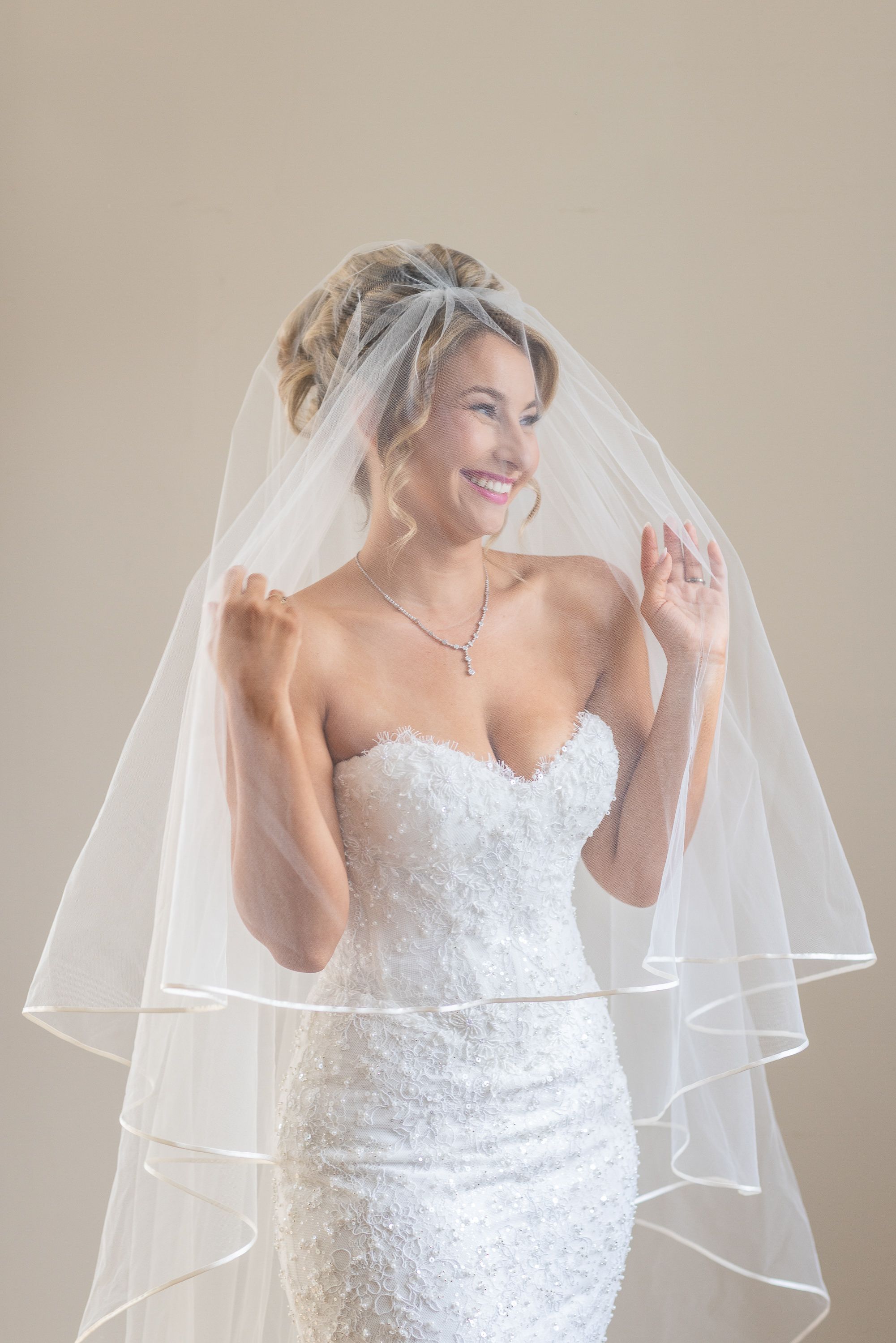 Smiling bride in strapless lace wedding gown and layered veil, holding veil open with both hands against neutral background.