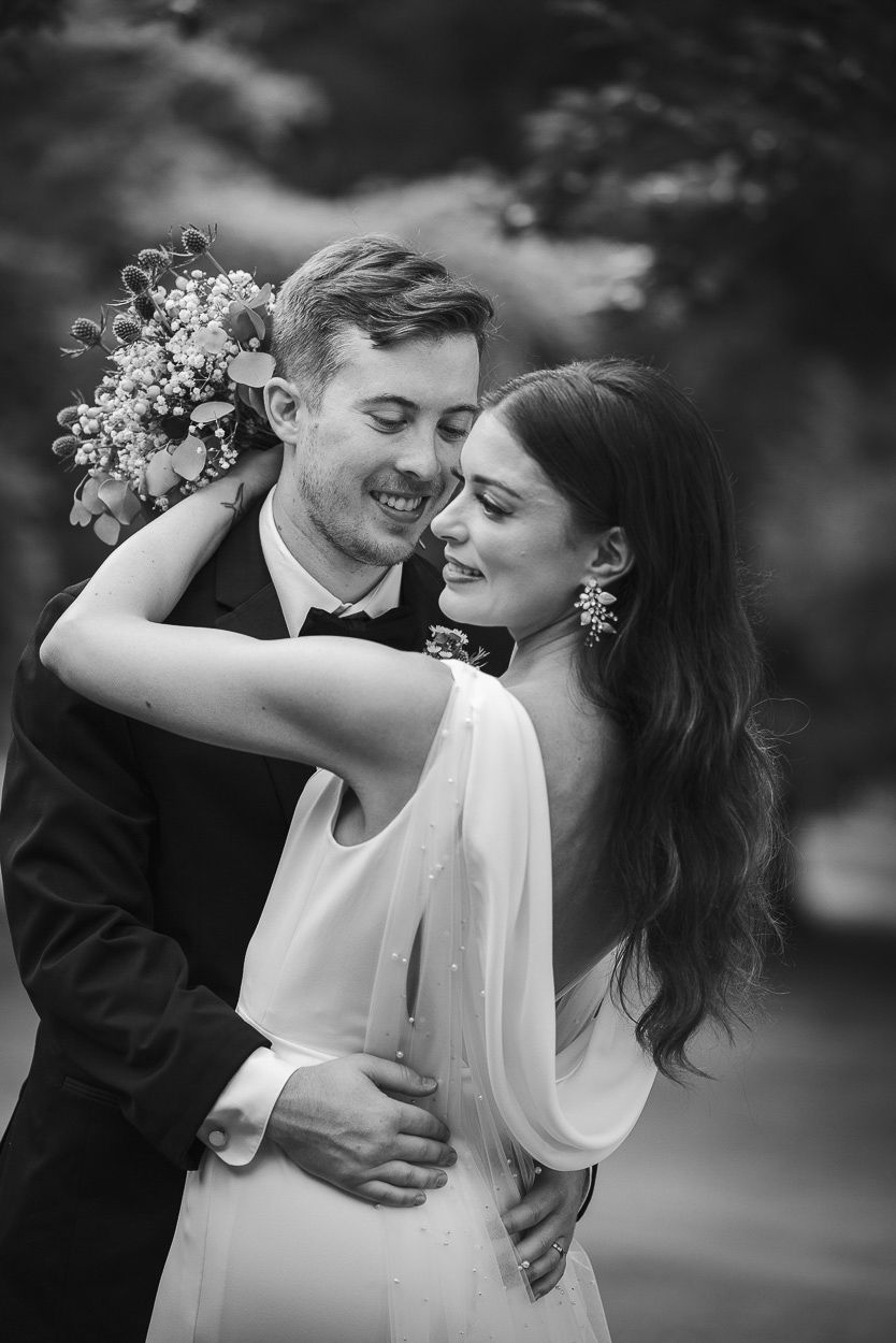 Black and white wedding photo of smiling bride and groom embracing outdoors, bride holding bouquet.
