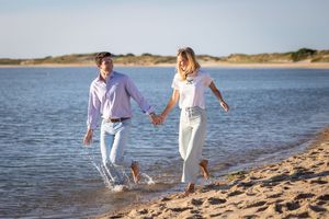 Couple holding hands walking barefoot along sandy beach shoreline with calm water and dunes in background.