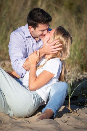Romantic couple kissing on sandy beach, man holding woman's face tenderly while sitting outdoors.