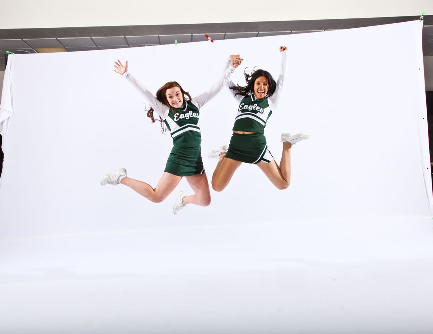 Two FHS cheerleaders wearing dark green uniforms jump high in the air against a white backdrop during a photo shoot.