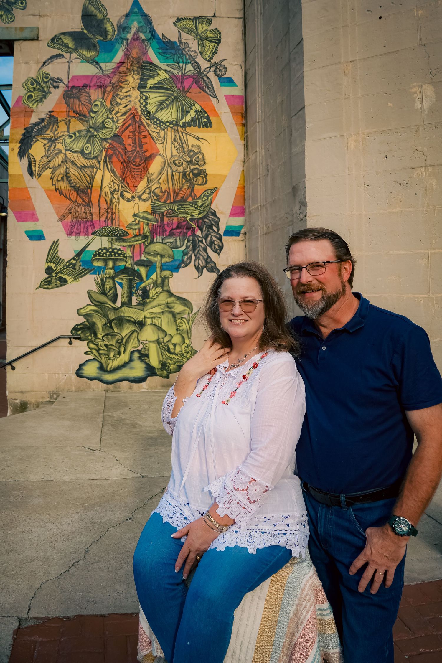 A couple poses in front of a vibrant botanical mural with green and yellow artwork on a beige wall.