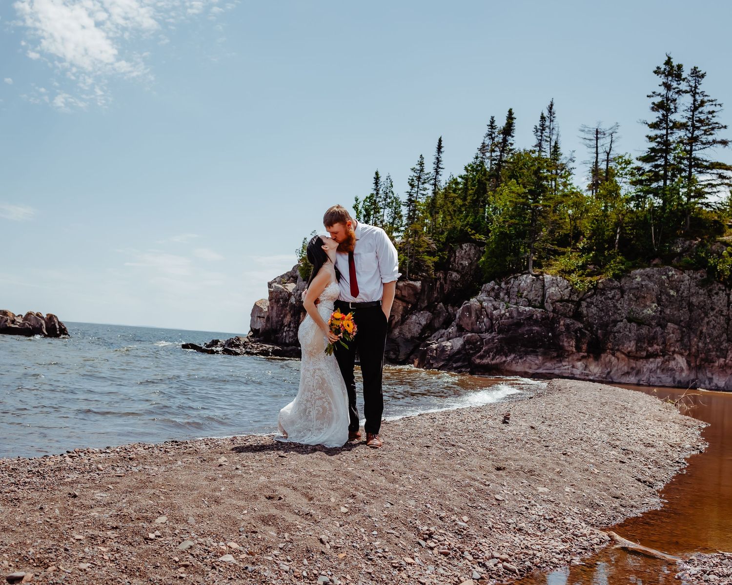 Wedding couple strolling along a scenic beach with pine-covered cliffs.