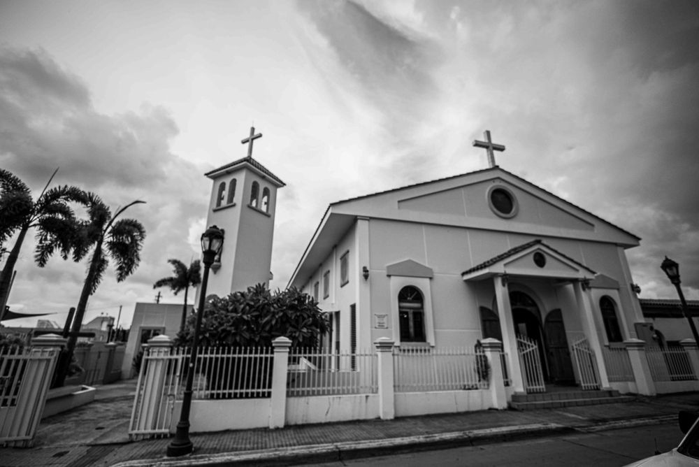 A white Catholic church exterior with bell tower and palm trees under dramatic cloudy skies.