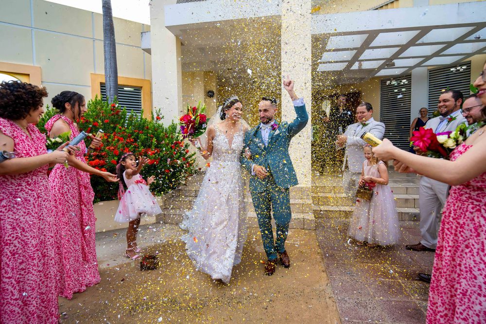 Wedding celebrants walk through gold confetti outside while guests cheer.