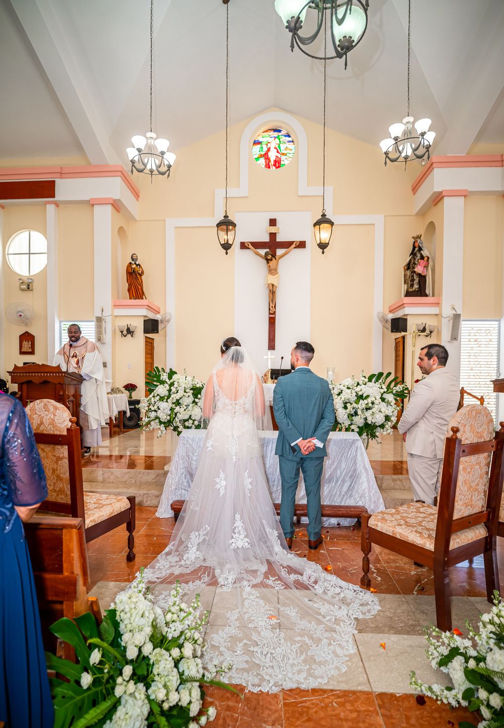 Church wedding ceremony with a long lace veil and white floral decorations beneath a hanging crucifix.