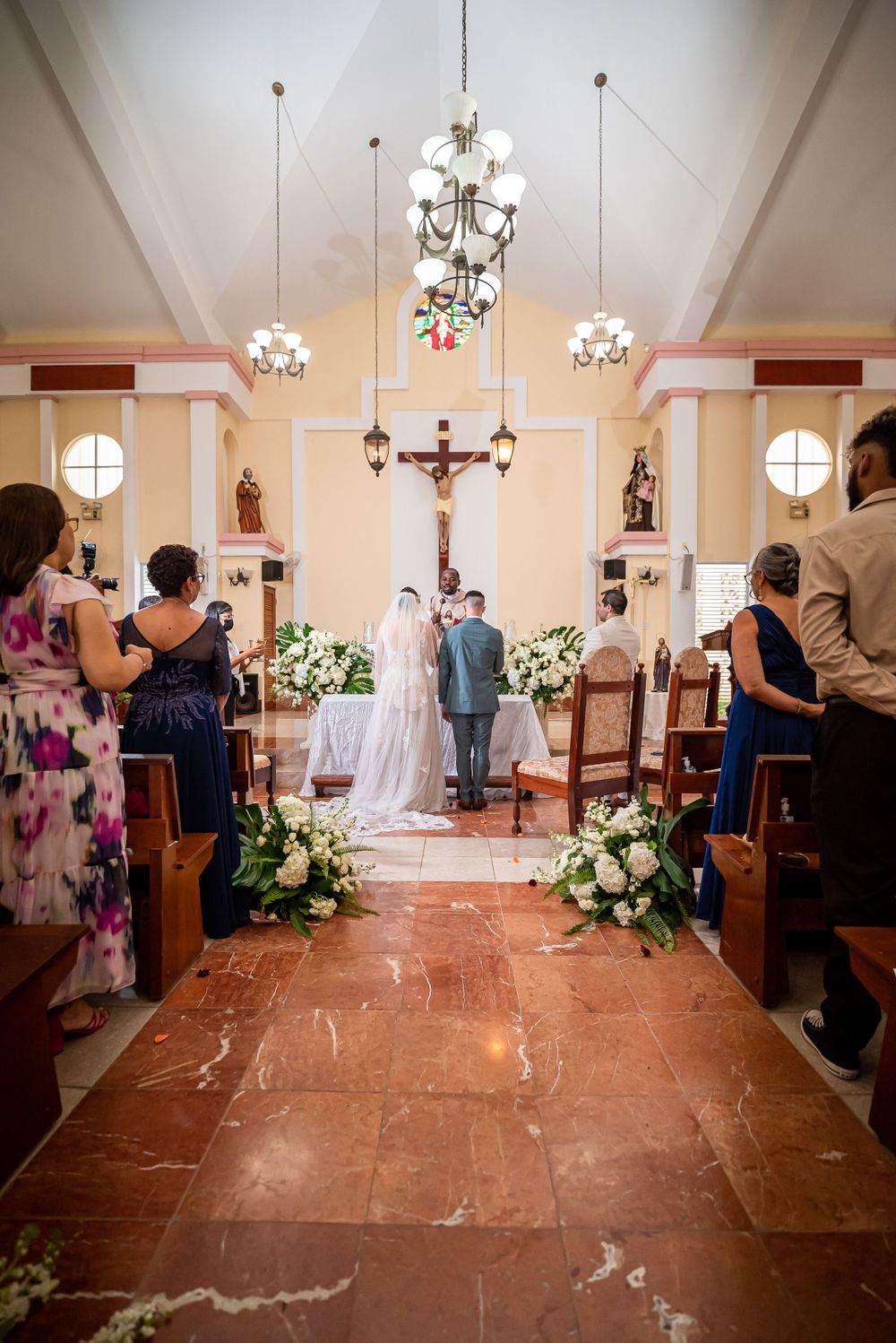 View down the church aisle with pendant lights and floral arrangements during a wedding ceremony.