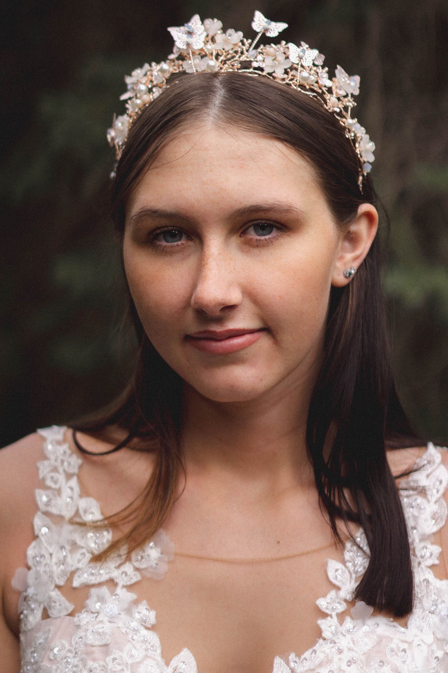 Young bride with brown hair wearing an ornate floral gold crown and lace wedding dress in forest.