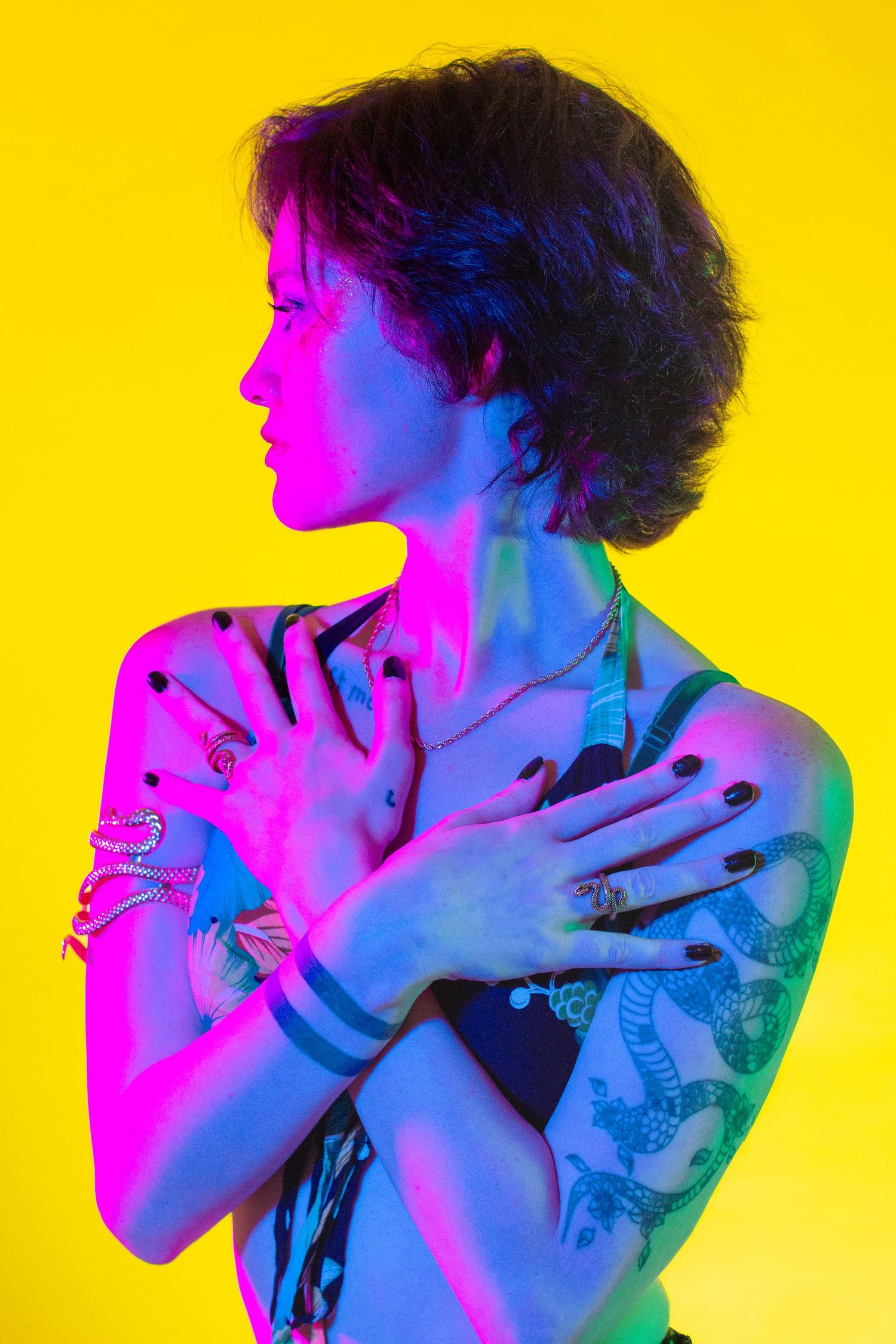 Artistic portrait with neon pink and blue lighting against yellow background showing tattoos and jewelry.