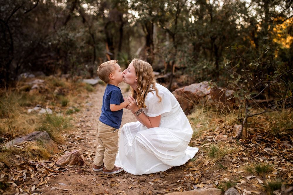Mother in white dress kisses young son on dirt trail surrounded by trees.