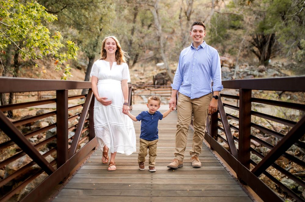 Pregnant woman in white dress with husband and toddler son posing on wooden bridge in nature.