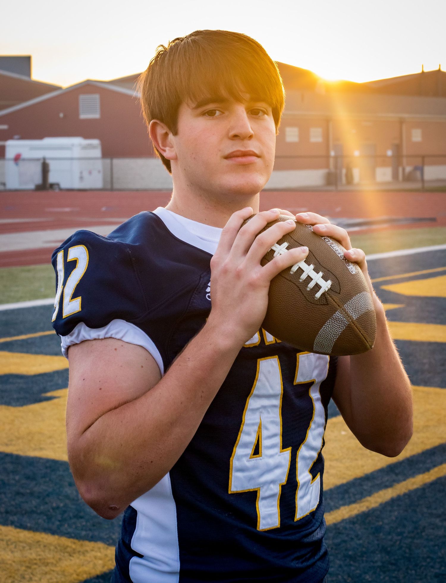 Football player in navy uniform number 42 holds ball during golden hour senior photo session.