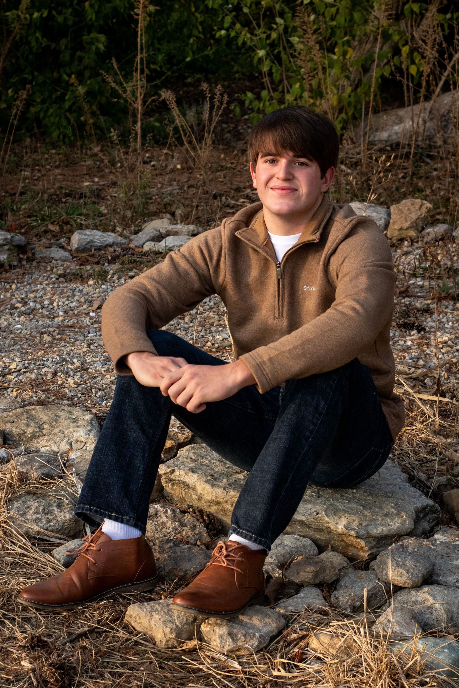 Senior portrait of student sitting on rocky ground wearing brown jacket and jeans.