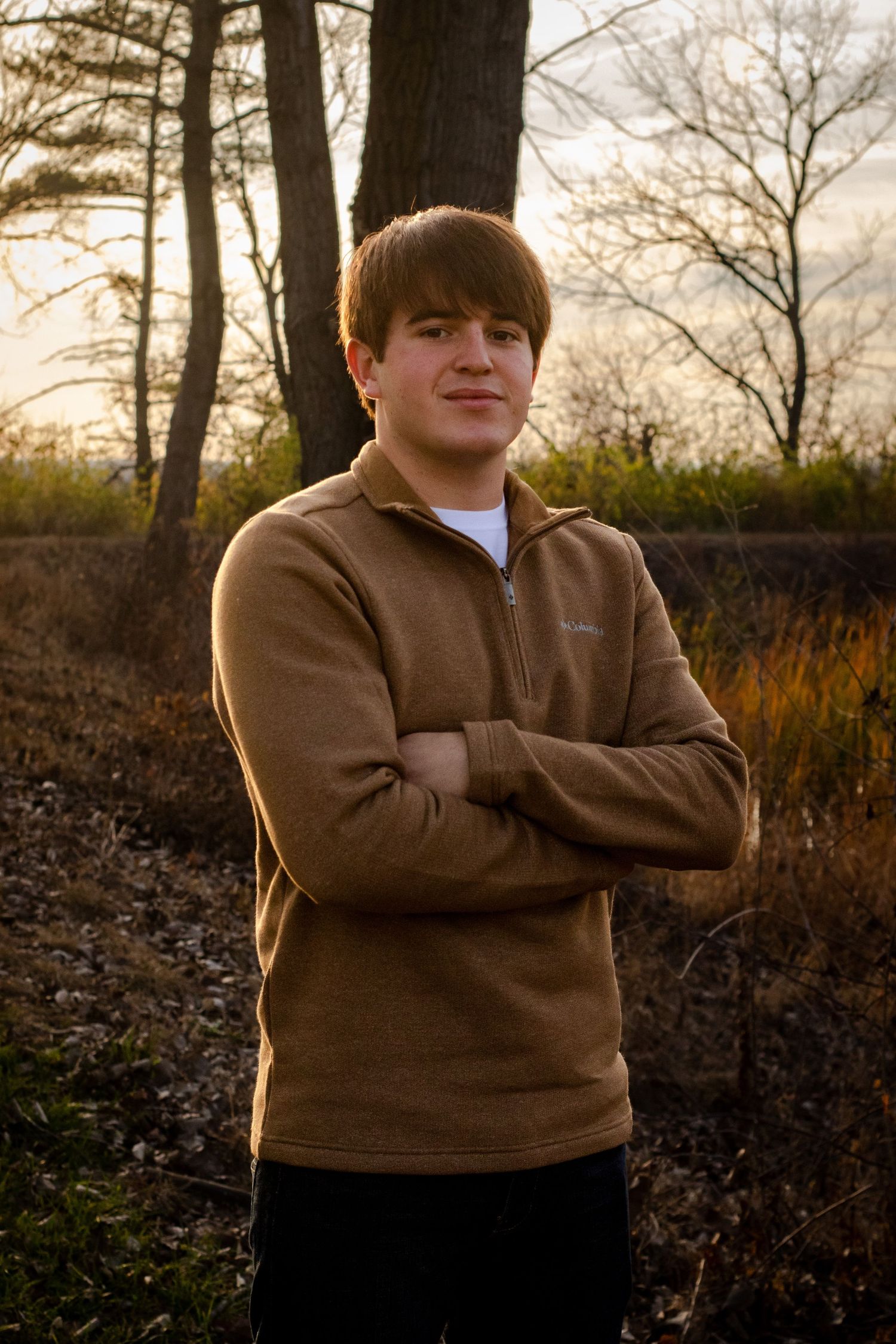 Student in brown pullover sweater poses outdoors at sunset.
