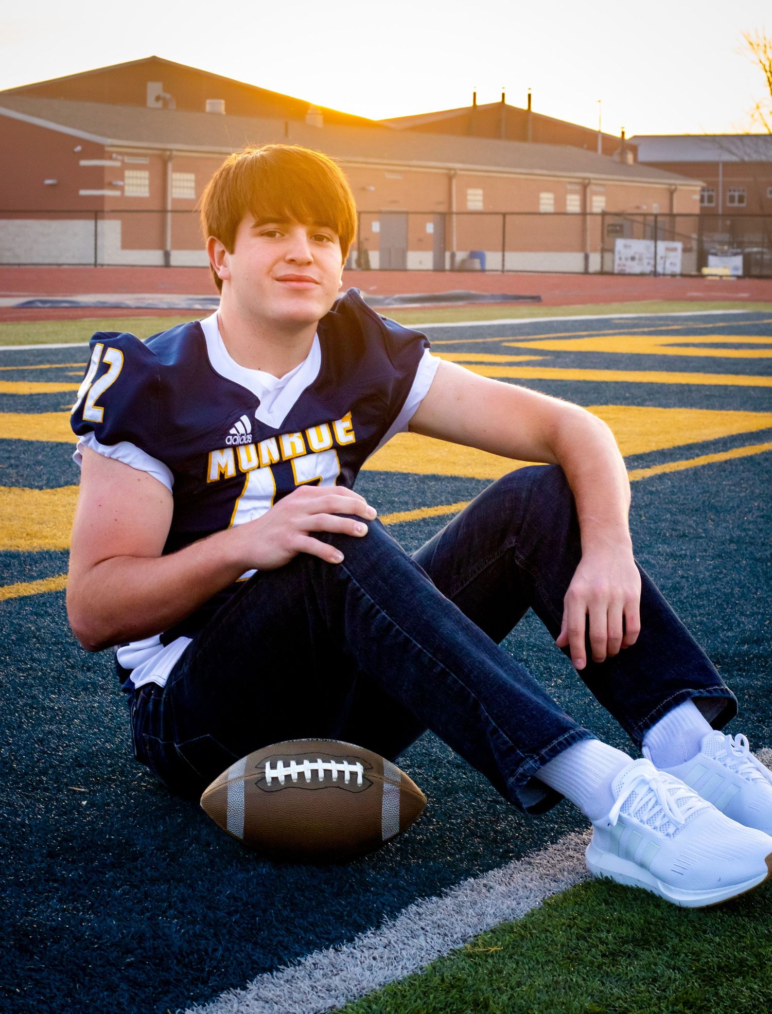 Football player in navy jersey posing with ball on field at sunset.