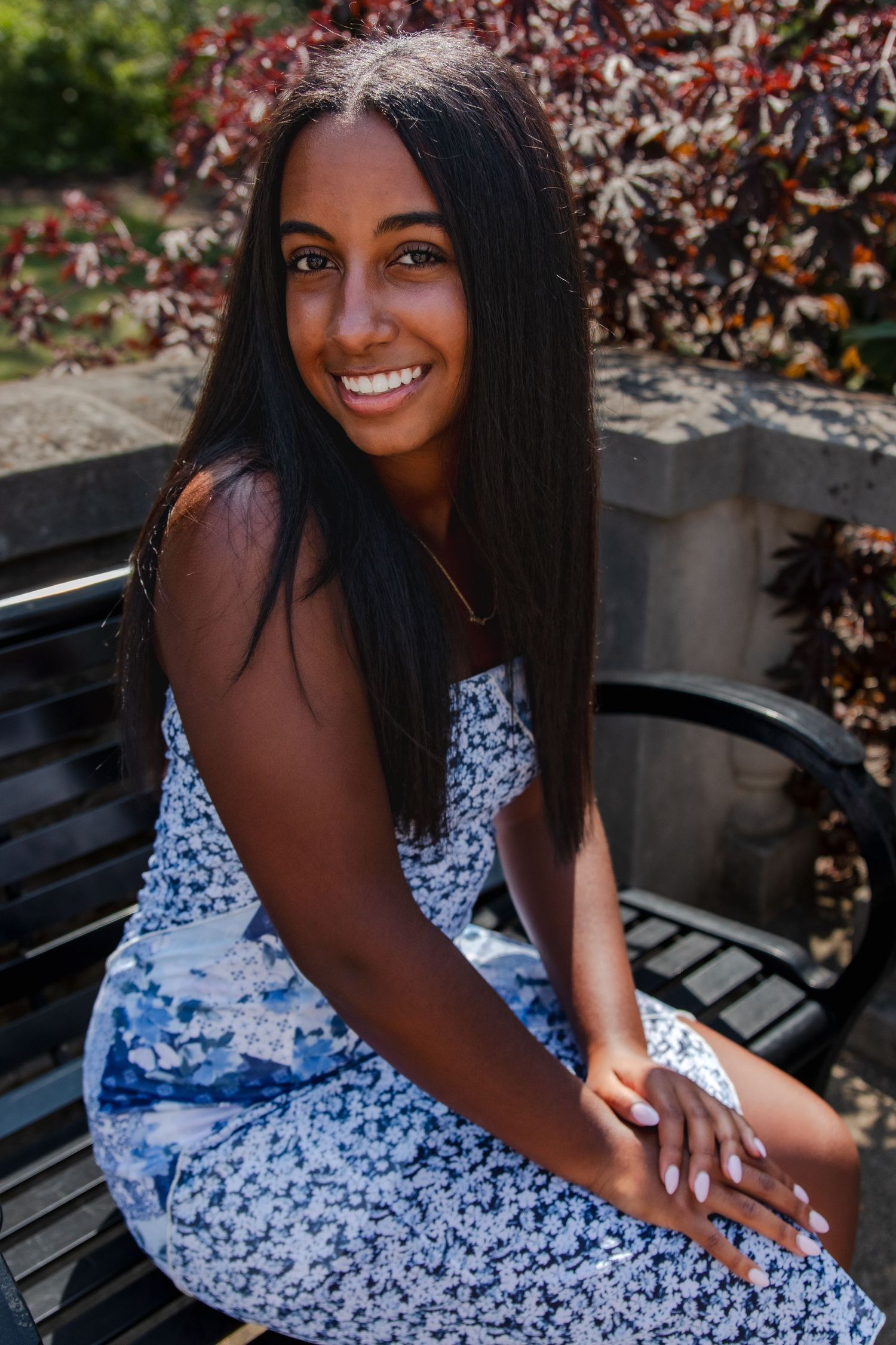 A senior in a blue and white floral dress poses gracefully on a wooden bench in a garden setting.