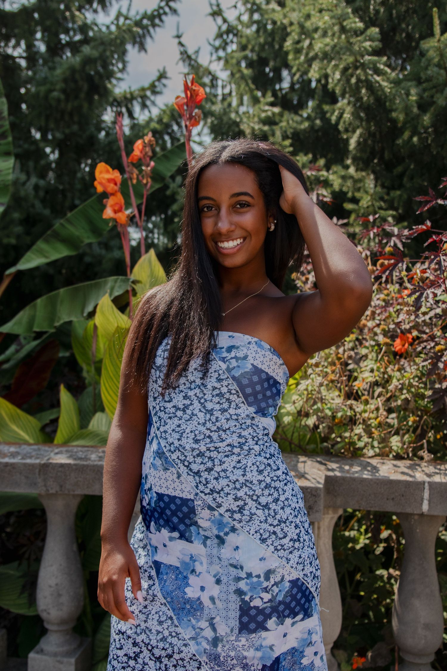 Senior in patterned blue dress smiles while posing in a garden setting with orange flowers.