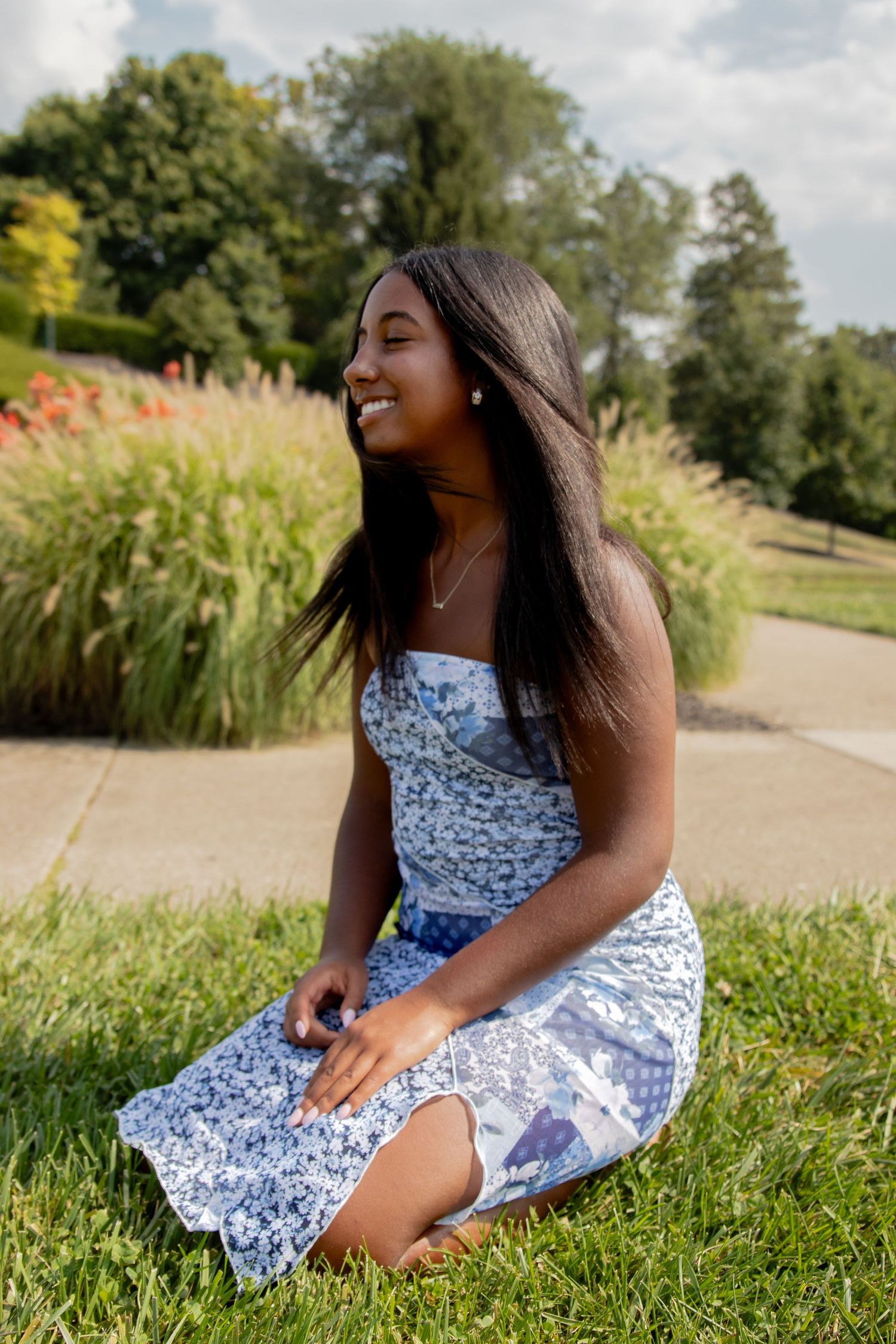 Senior in floral dress sits gracefully on grass during a sunny outdoor photoshoot.