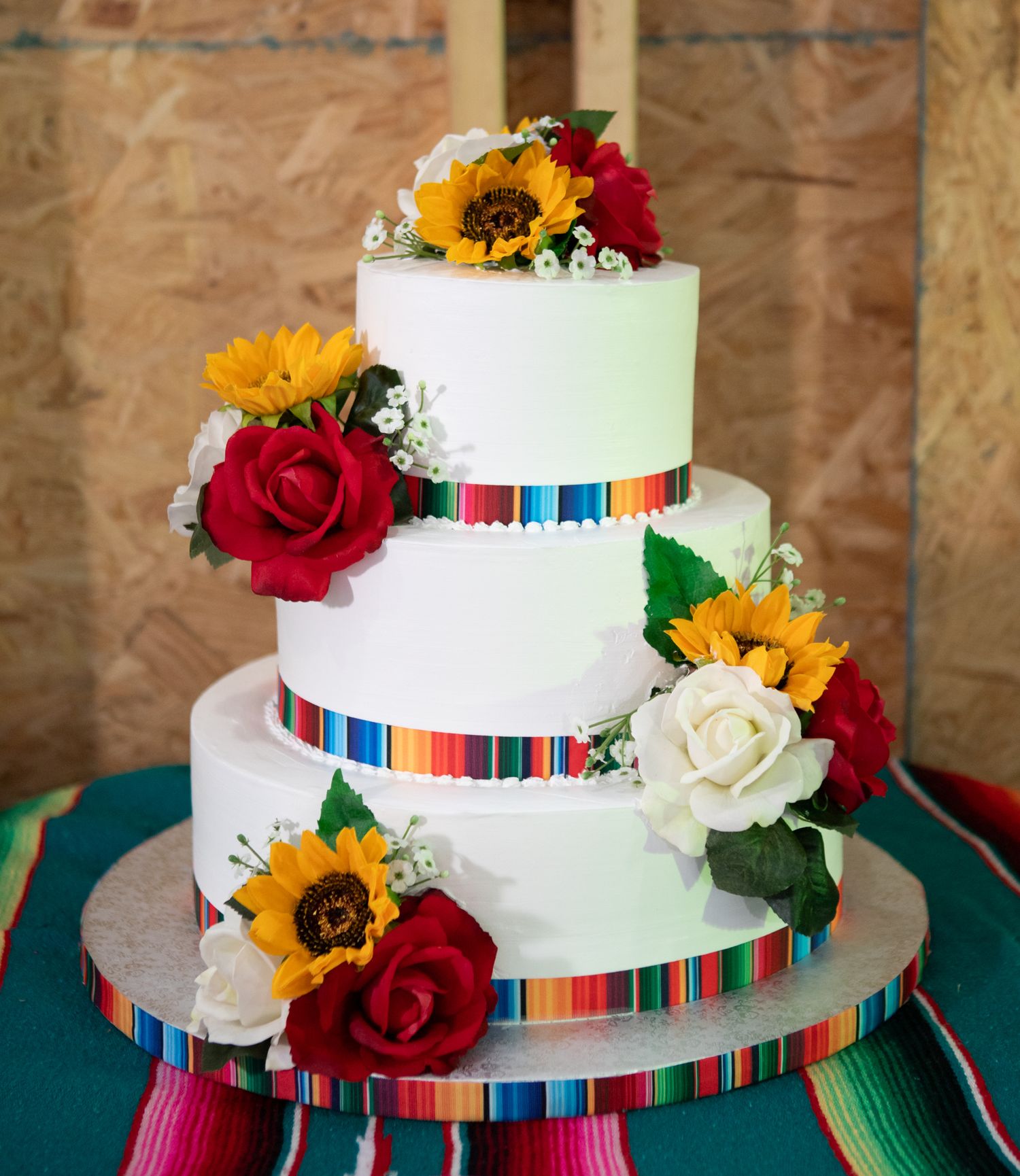 White tiered wedding cake decorated with colorful serape ribbons, sunflowers, and red roses.