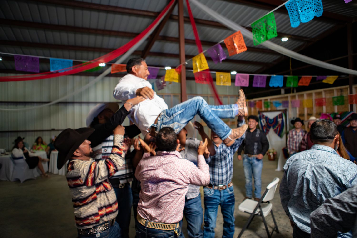 Wedding guests toss someone into the air during a festive celebration with papel picado decorations.