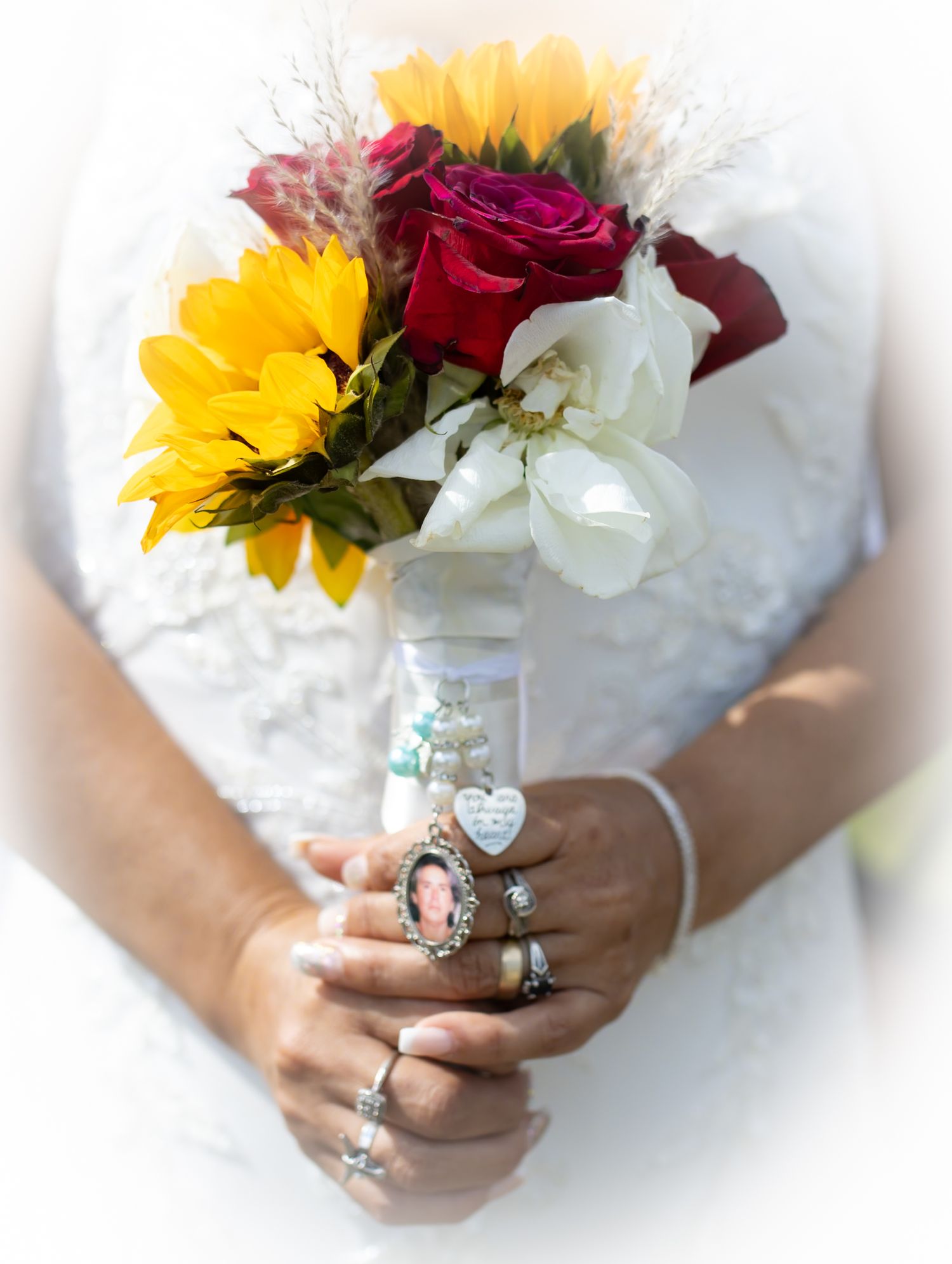Wedding bouquet with sunflowers, red roses and white flowers held by hands adorned with rings.