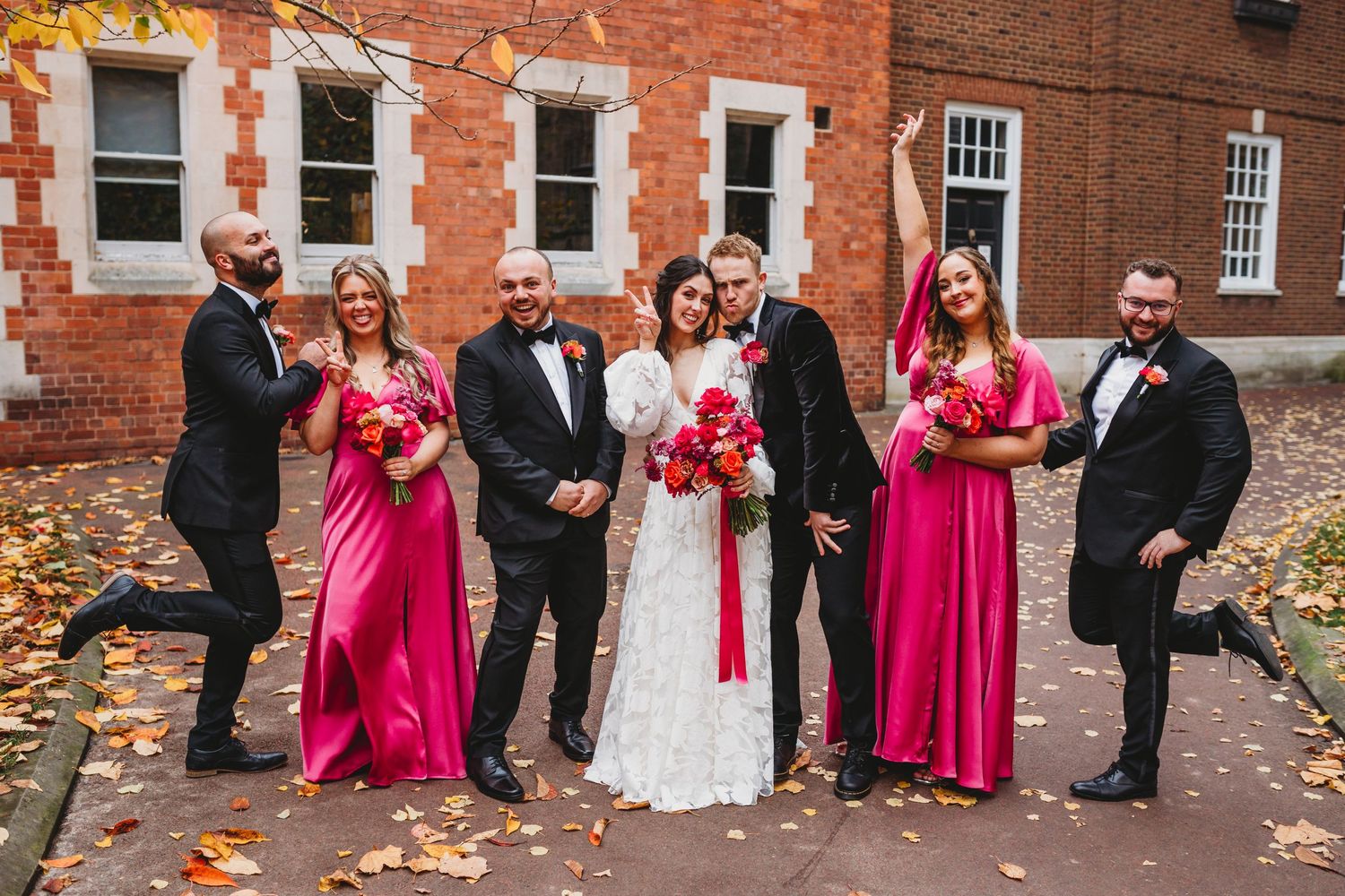 Wedding party in bright pink dresses poses against brick building in autumn.