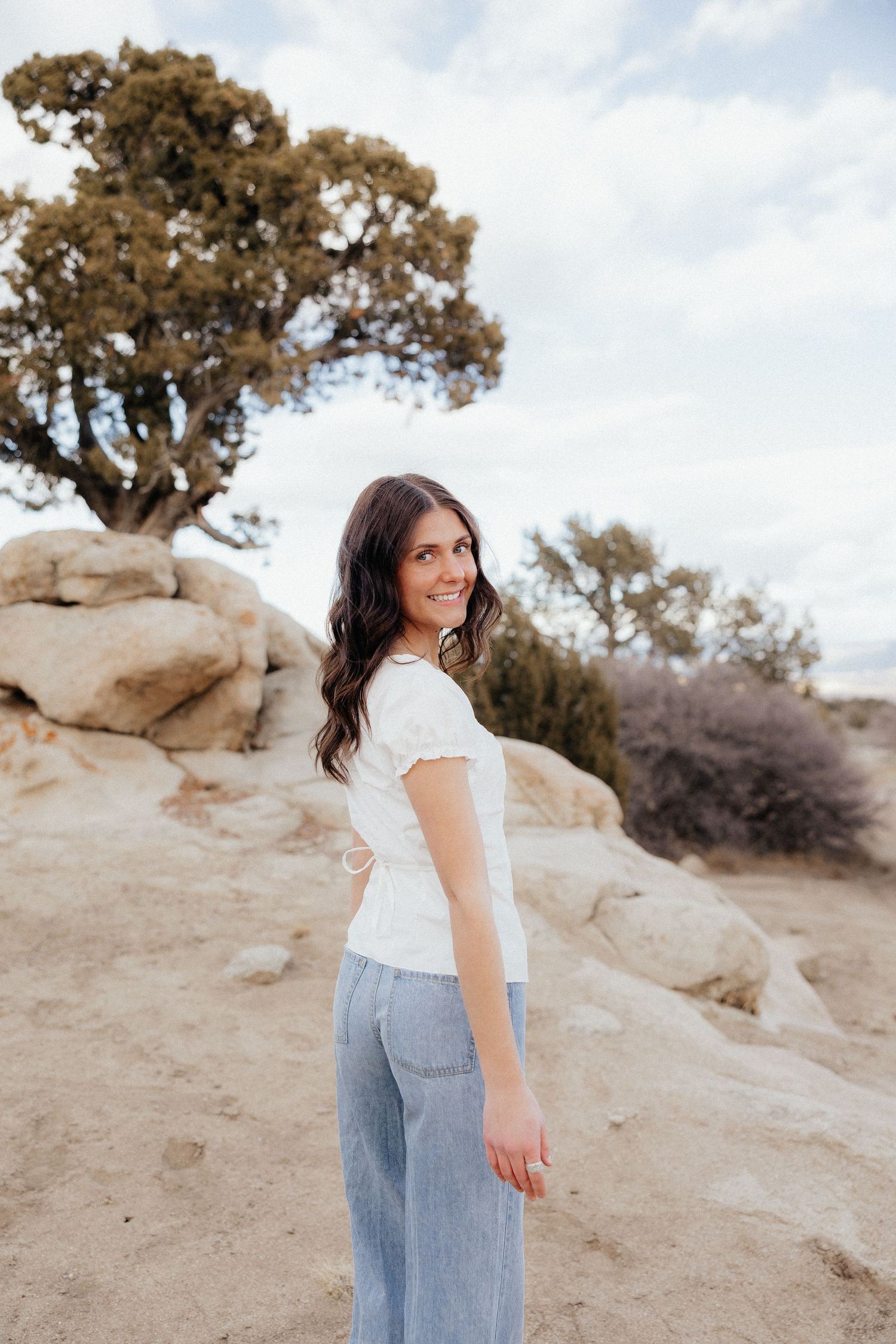 Person in white t-shirt and jeans stands on sandy terrain with a tree against cloudy sky background in natural setting.