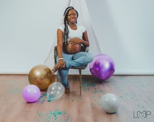Person sitting on floor surrounded by colorful balloons and confetti in a minimalist white room setting.