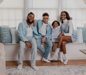 Family wearing matching light blue pajamas sits together at home on a cozy white window bench.