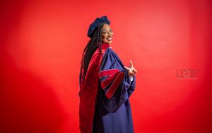 Graduate in blue academic regalia laughs joyfully against vibrant red background during graduation photoshoot.