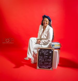 Graduate in white regalia and blue cap sits on cube with chalkboard against vibrant red background during photo shoot.